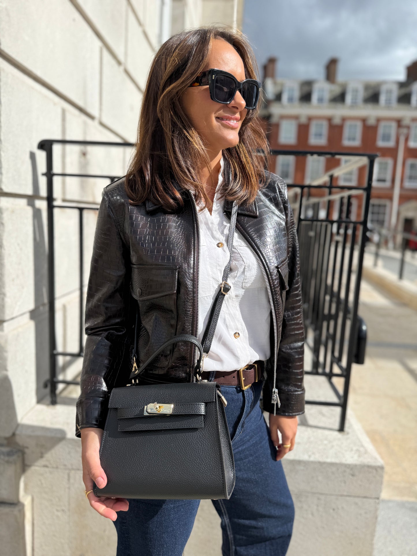 Woman in a black leather jacket and sunglasses holding a black handbag on a city street.