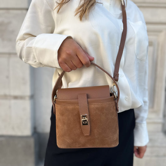 A person wearing a white shirt and  brown suede personalized leather crossbody bag with a gold hardware twist lock closure.