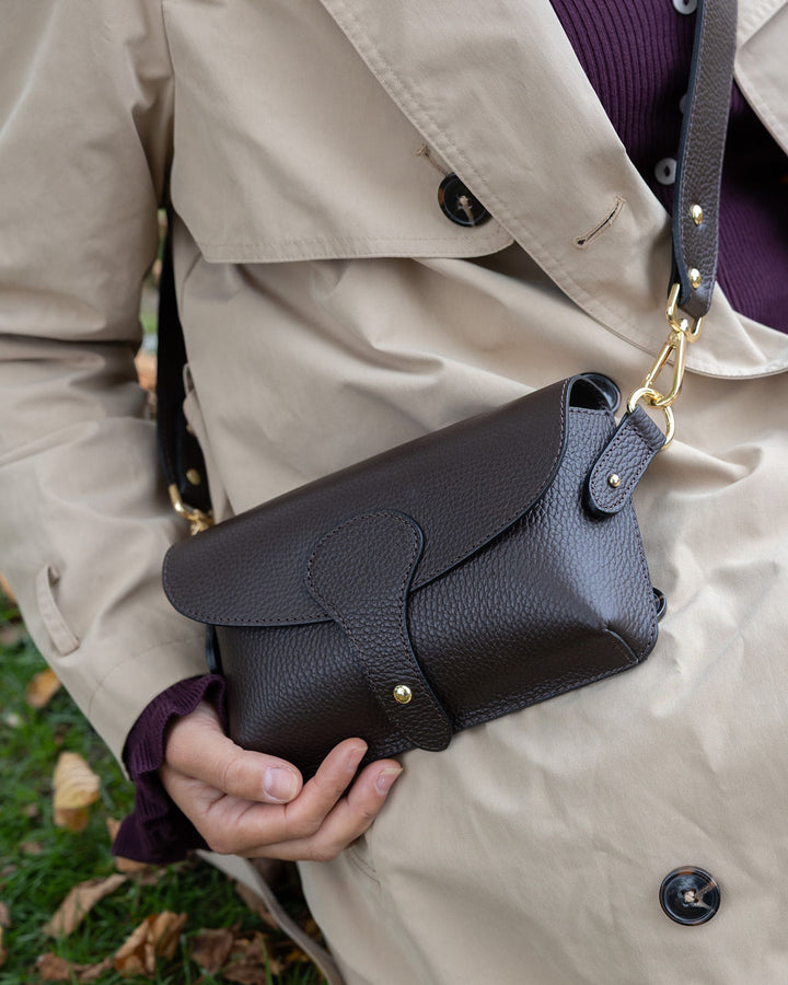 Person holding a dark brown  leather handbag with a gold clasp outdoors.