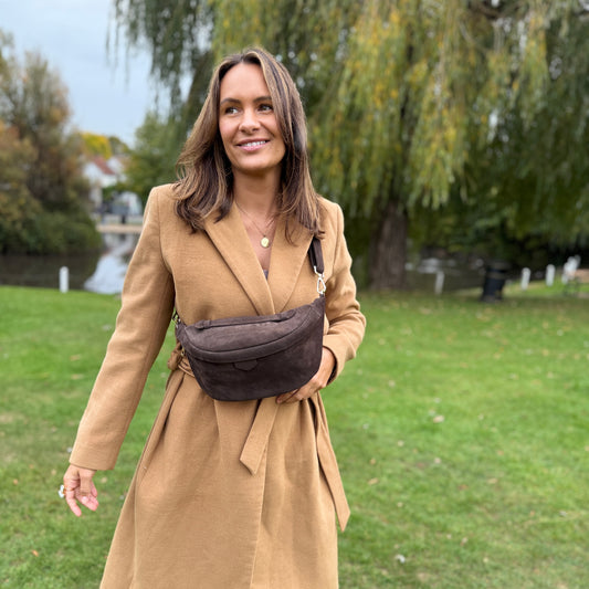 Woman in a beige coat with a brown suede waist bag standing in a park.