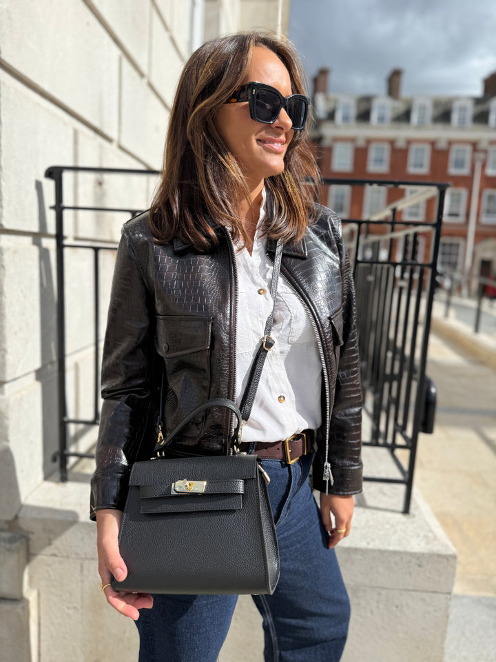 Woman in a black leather jacket and sunglasses holding a black leather handbag on a city street.