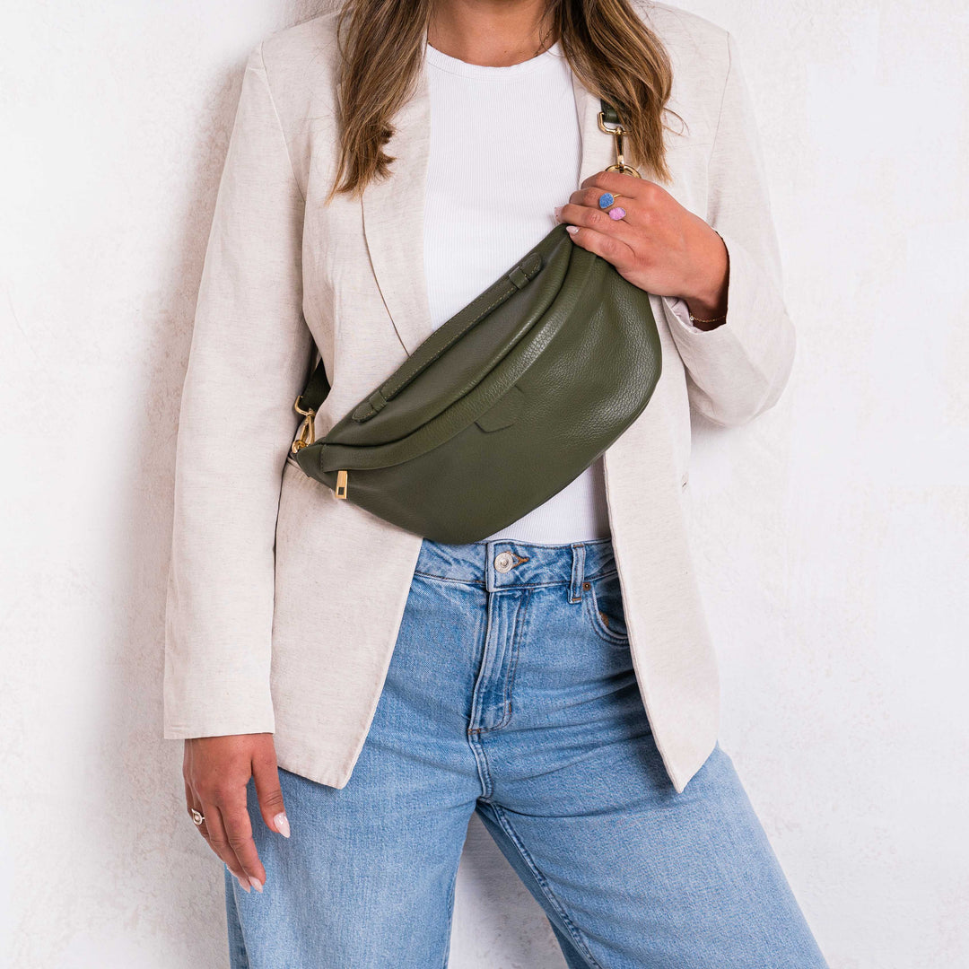 A person wearing a beige jacket and blue jeans is holding a Khaki green leather bumbag waist bag with a gold zipper.