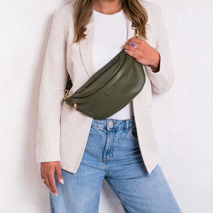 A person wearing a beige jacket and blue jeans is holding a Khaki green leather bumbag waist bag with a gold zipper.