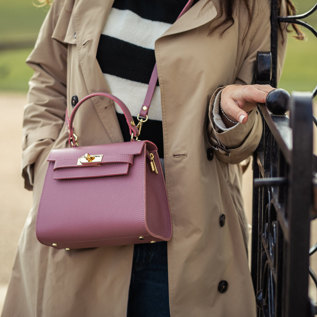 A person wearing a beige coat is holding a brown leather top handle crossbody bag with a gold turn lock closure.