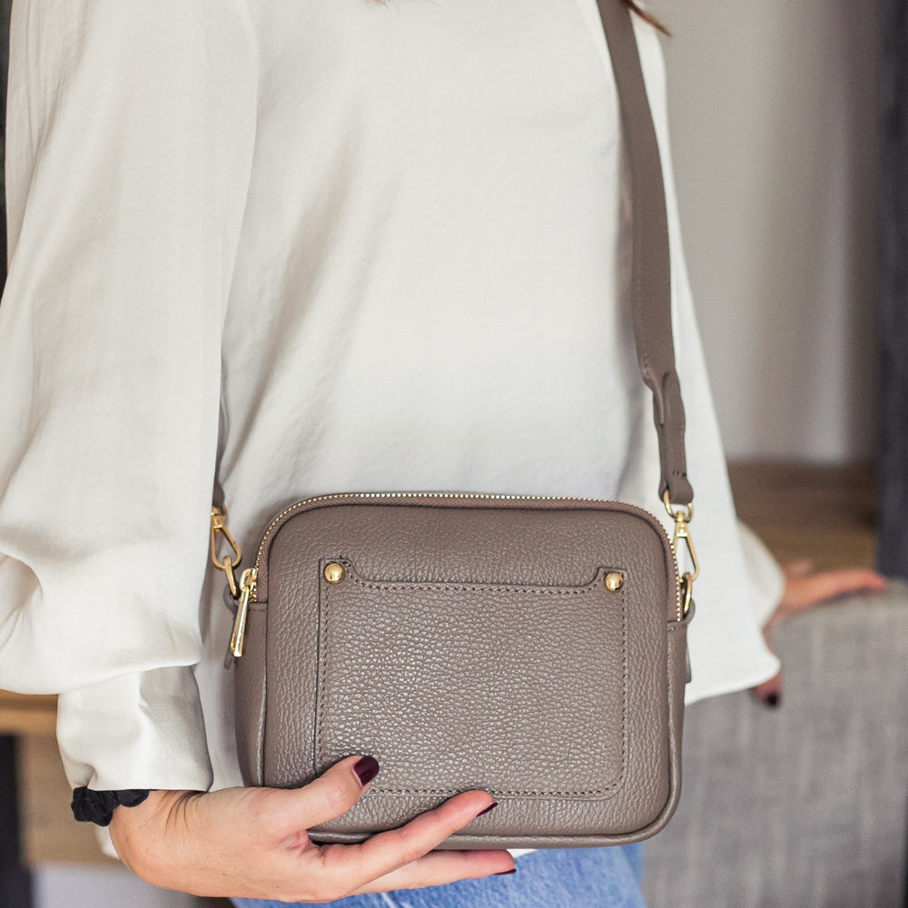 A woman carrying a dark taupe leather double zip crossbody bag with a detachable strap over her shoulder.