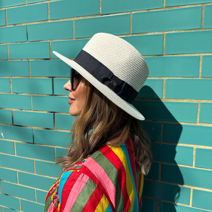 Woman wearing a cream straw fedora hat with a black band with personalised initials against a teal brick wall.