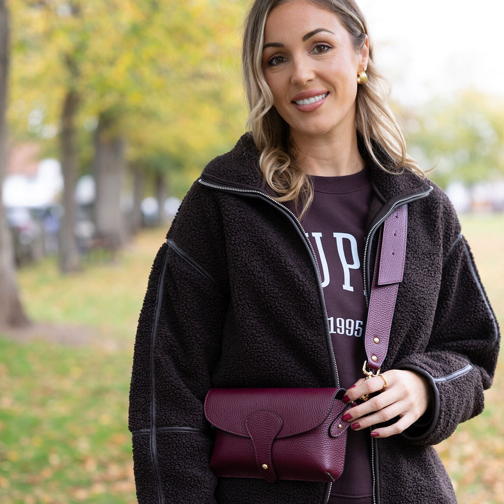 Woman holding a burgundy handbag in an outdoor setting with trees and grass.