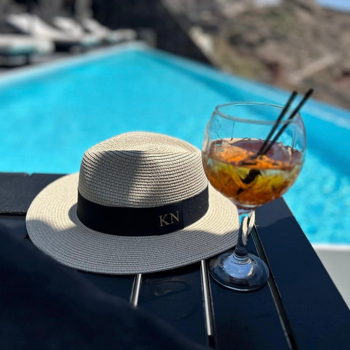 Straw hat with a black band and initials next to a cocktail by a pool